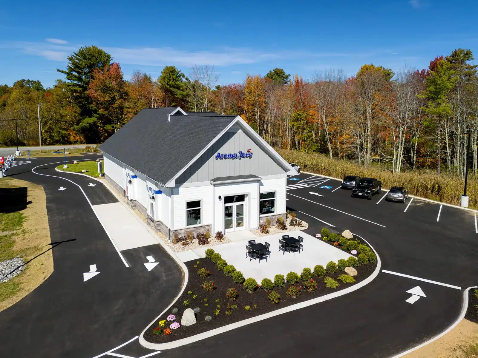 Aerial view of an Aroma Joe’s coffee shop with drive-thru lanes, outdoor seating, landscaped island, and surrounding parking lot.
